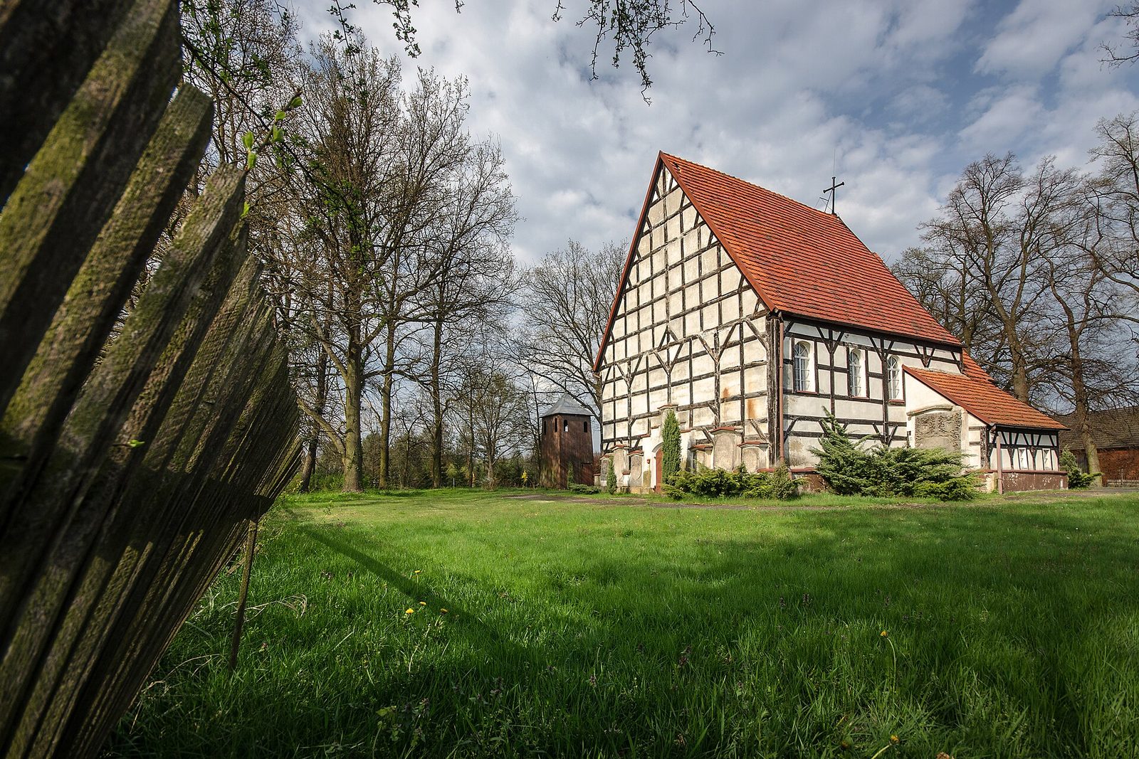 Szachulcowy kościół Matki Bożej Różańcowej w Trzmielowie z czerwonym dachem, w tle dzwonnica i drzewa