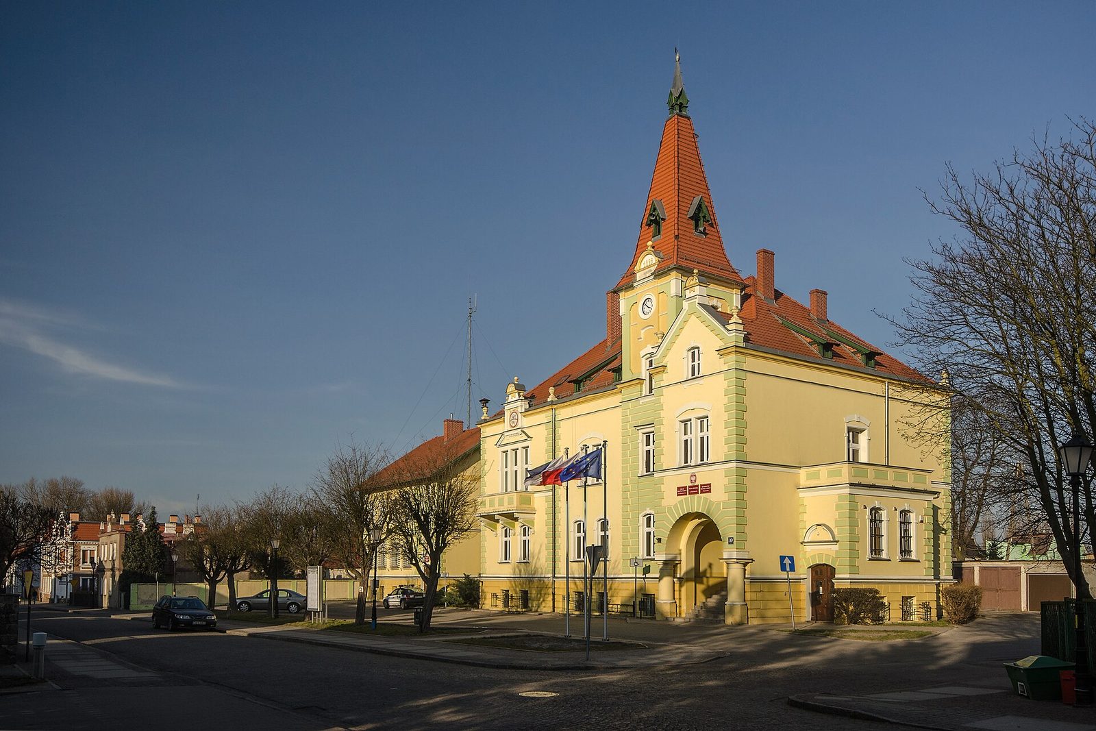 Żółty ratusz w Chocianowie z 1899 roku z wieżą zegarową krytą czerwoną dachówką i flagami przy wejściu